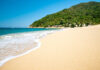 A pristine, empty beach with gentle waves coming in from the Pacific Ocean in Yelapa, Jalisco. In the background is a palm-tree-covered mountain.