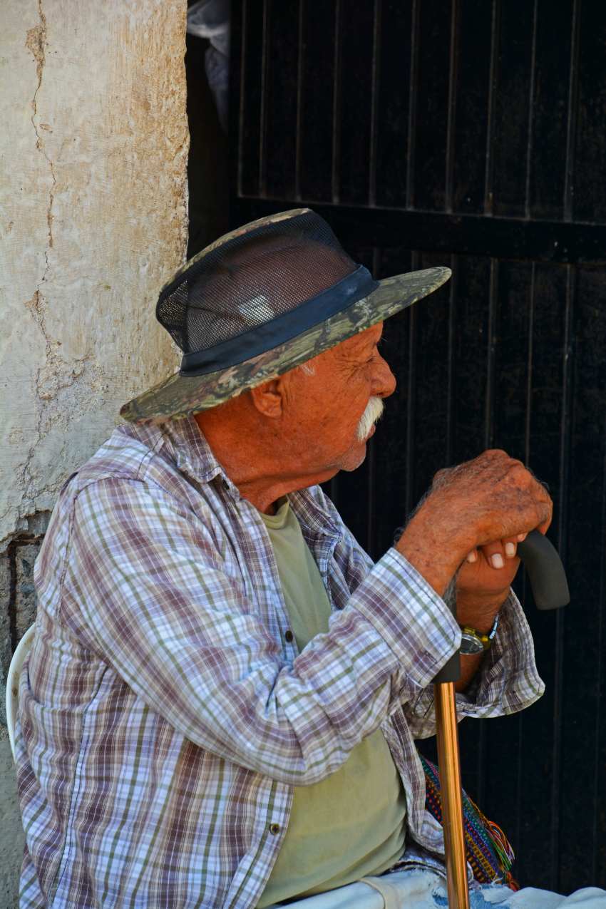 An elderly man sitting with a cane outside an adobe building on a Mexican street