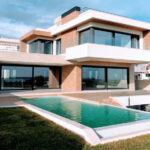 A bright, full exterior shot showcases a large, modern luxury home with a rectangular infinity pool in the foreground. The house features a striking contemporary design with white and earth-toned facades, expansive dark-framed windows and sliding glass doors, and multiple levels. A grassy lawn surrounds the pool, and in the distance, other residential buildings and clear skies with scattered clouds are visible.