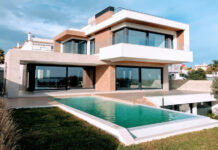 A bright, full exterior shot showcases a large, modern luxury home with a rectangular infinity pool in the foreground. The house features a striking contemporary design with white and earth-toned facades, expansive dark-framed windows and sliding glass doors, and multiple levels. A grassy lawn surrounds the pool, and in the distance, other residential buildings and clear skies with scattered clouds are visible.