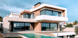 A bright, full exterior shot showcases a large, modern luxury home with a rectangular infinity pool in the foreground. The house features a striking contemporary design with white and earth-toned facades, expansive dark-framed windows and sliding glass doors, and multiple levels. A grassy lawn surrounds the pool, and in the distance, other residential buildings and clear skies with scattered clouds are visible.