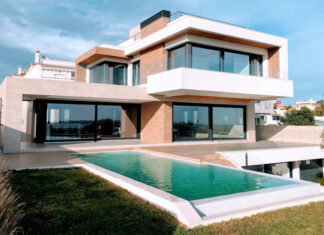 A bright, full exterior shot showcases a large, modern luxury home with a rectangular infinity pool in the foreground. The house features a striking contemporary design with white and earth-toned facades, expansive dark-framed windows and sliding glass doors, and multiple levels. A grassy lawn surrounds the pool, and in the distance, other residential buildings and clear skies with scattered clouds are visible.