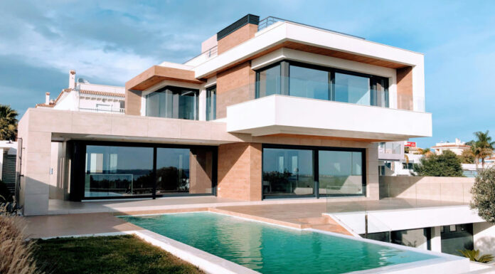 A bright, full exterior shot showcases a large, modern luxury home with a rectangular infinity pool in the foreground. The house features a striking contemporary design with white and earth-toned facades, expansive dark-framed windows and sliding glass doors, and multiple levels. A grassy lawn surrounds the pool, and in the distance, other residential buildings and clear skies with scattered clouds are visible.