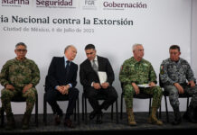 Security chief Omar García Harfuch, Attorney General Gertz and other Mexian officials sit on a stage in front of a banner reading "National Strategy against Extortion" in spanish