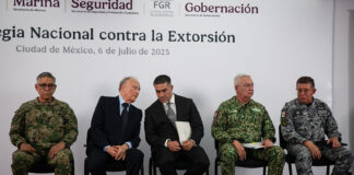 Security chief Omar García Harfuch, Attorney General Gertz and other Mexian officials sit on a stage in front of a banner reading "National Strategy against Extortion" in spanish