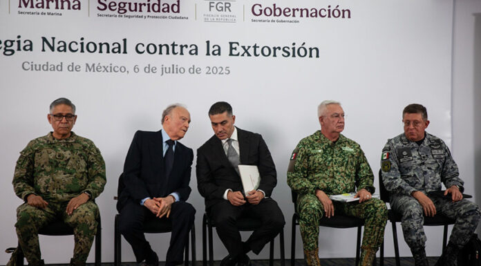 Security chief Omar García Harfuch, Attorney General Gertz and other Mexian officials sit on a stage in front of a banner reading "National Strategy against Extortion" in spanish