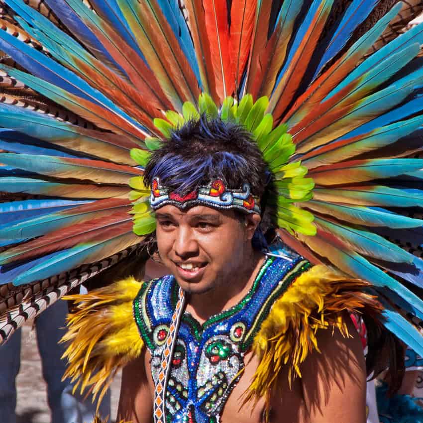A smiling traditional Mexican dancer wearing an elaborate headdress of vibrant blue, orange, and green feathers, with a matching embroidered vest and yellow feathered shoulders.