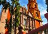 Close-up of a historic church in San Miguel de Allende, showcasing its distinctive orange and yellow colors, ornate facade, and bell tower, with a cypress tree in the foreground.
