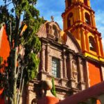 Close-up of a historic church in San Miguel de Allende, showcasing its distinctive orange and yellow colors, ornate facade, and bell tower, with a cypress tree in the foreground.