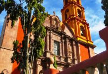 Close-up of a historic church in San Miguel de Allende, showcasing its distinctive orange and yellow colors, ornate facade, and bell tower, with a cypress tree in the foreground.