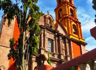 Close-up of a historic church in San Miguel de Allende, showcasing its distinctive orange and yellow colors, ornate facade, and bell tower, with a cypress tree in the foreground.