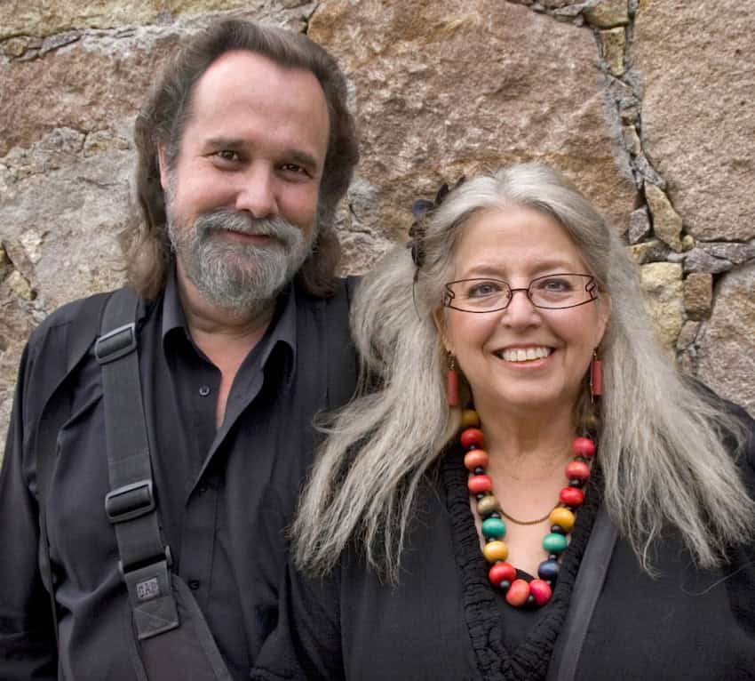 Cathi and Steven House, the authors of the San Miguel de Allende photography book, smile in front of a stone wall. Steven has a beard and long dark hair, and Cathi has long gray hair and is wearing a colorful beaded necklace.