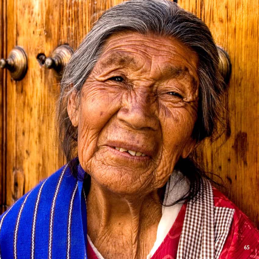 A close-up portrait of an elderly Mexican woman with warm, crinkled eyes and a gentle smile, wearing a blue and white striped rebozo over a red and checkered blouse, against a rustic wooden door.