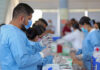 A Mexican man in scrubs and a facemask fills a syringe from a vial in a busy vaccine application center