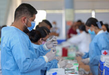 A Mexican man in scrubs and a facemask fills a syringe from a vial in a busy vaccine application center