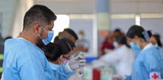 A Mexican man in scrubs and a facemask fills a syringe from a vial in a busy vaccine application center