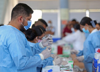 A Mexican man in scrubs and a facemask fills a syringe from a vial in a busy vaccine application center