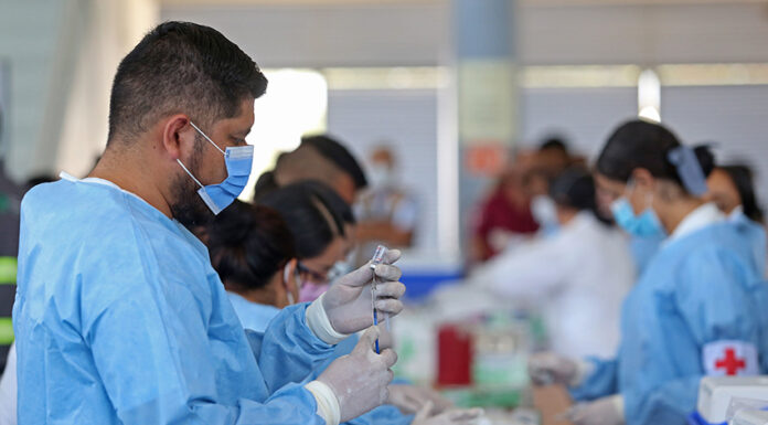 A Mexican man in scrubs and a facemask fills a syringe from a vial in a busy vaccine application center