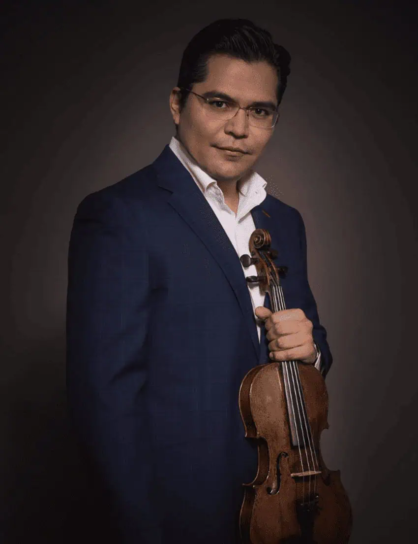 A portrait-style photo features a Mexican violonist standing against a dark, plain background. He is wearing a dark blue suit jacket over a white collared shirt, open at the neck, and clear-framed glasses. He holds a violin in his right hand, resting it against his chest. He looks directly at the viewer with a neutral expression.