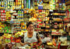 A Mexican middle-aged woman with a warm smile stands behind a counter laden with colorful packaged goods in her small, traditional Mexican grocery store. Shelves packed tightly with a wide variety of canned goods, bottles, and boxes rise behind her create a vibrant, somewhat cluttered backdrop. She wears a white apron dress over her sundress to protect it.
