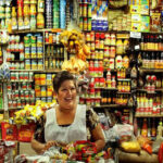 A Mexican middle-aged woman with a warm smile stands behind a counter laden with colorful packaged goods in her small, traditional Mexican grocery store. Shelves packed tightly with a wide variety of canned goods, bottles, and boxes rise behind her create a vibrant, somewhat cluttered backdrop. She wears a white apron dress over her sundress to protect it.