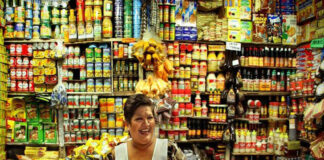 A Mexican middle-aged woman with a warm smile stands behind a counter laden with colorful packaged goods in her small, traditional Mexican grocery store. Shelves packed tightly with a wide variety of canned goods, bottles, and boxes rise behind her create a vibrant, somewhat cluttered backdrop. She wears a white apron dress over her sundress to protect it.