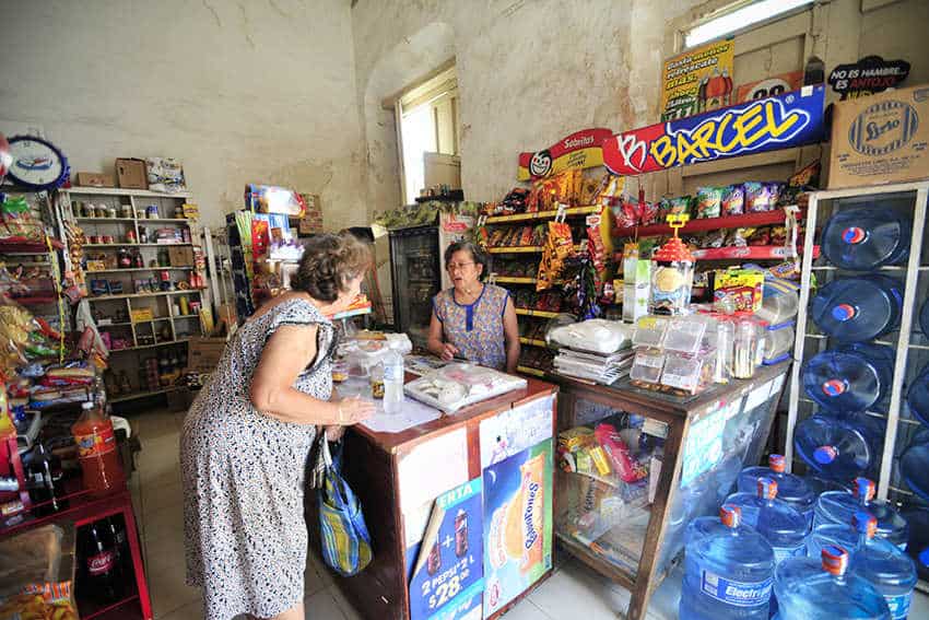 In a rustic, old-fashioned Mexican "tiendita" or small grocery store, two women are engaged in a transaction. One woman, with her back mostly to the viewer, is leaning over a wooden counter, appearing to be a customer. Facing her, from behind the counter, is another woman who seems to be the shopkeeper. The store is packed with various goods, including shelves filled with packaged items, bottled water jugs, and colorful snack advertisements.