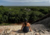 A person with their back to the camera sits on a stone edge, looking out over a vast, dense green jungle canopy that stretches to the horizon under a cloudy sky. In the distance, ruins are partially visible above the treetops.
