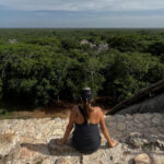 A person with their back to the camera sits on a stone edge, looking out over a vast, dense green jungle canopy that stretches to the horizon under a cloudy sky. In the distance, ruins are partially visible above the treetops.