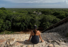A person with their back to the camera sits on a stone edge, looking out over a vast, dense green jungle canopy that stretches to the horizon under a cloudy sky. In the distance, ruins are partially visible above the treetops.