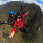 A woman in a red jacket smiles widely while tandem paragliding over a mountainous landscape with a professional pilot.