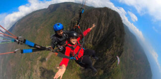 A woman in a red jacket smiles widely while tandem paragliding over a mountainous landscape with a professional pilot.
