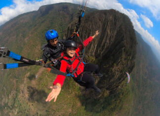 A woman in a red jacket smiles widely while tandem paragliding over a mountainous landscape with a professional pilot.
