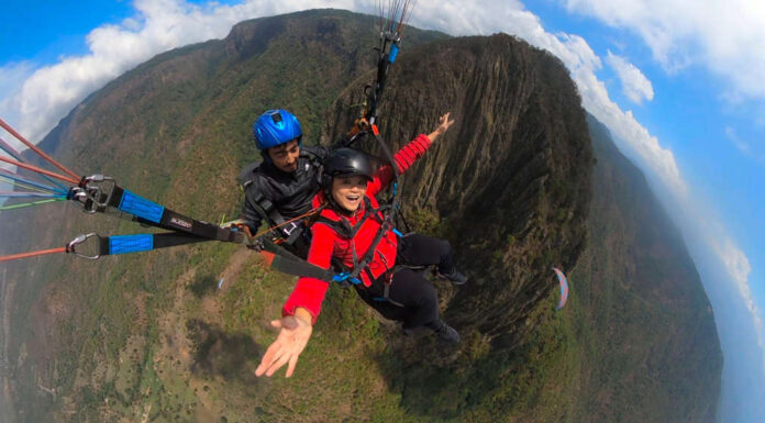 A woman in a red jacket smiles widely while tandem paragliding over a mountainous landscape with a professional pilot.