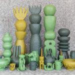 A collection of unique, handcrafted Mexican ceramic art pieces in shades of green and yellow, including tall stacked sculptures, vases, bowls, and whimsical figures, arranged on a textured grey surface.