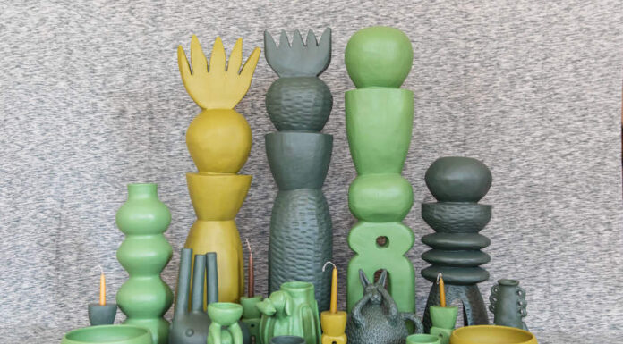 A collection of unique, handcrafted Mexican ceramic art pieces in shades of green and yellow, including tall stacked sculptures, vases, bowls, and whimsical figures, arranged on a textured grey surface.