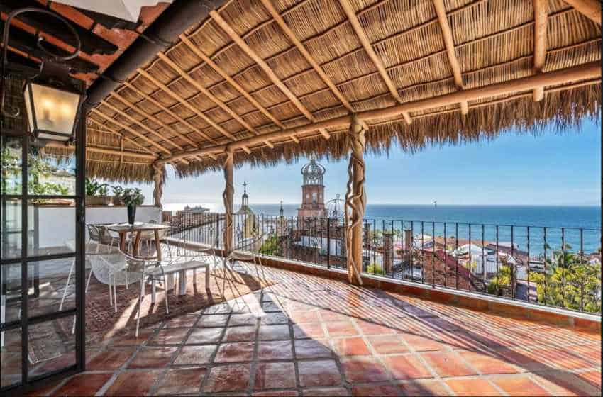 A spacious outdoor rooftop patio in a Puerto Vallarta home has a thatched roof and overlooks the ocean and a city, all under a clear blue sky.