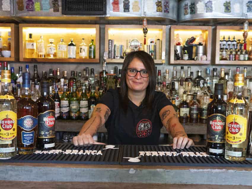 Un sourire avec des mains et des miroirs tatoués, Claudia Caprera, se tient derrière un bar rempli de bouteilles d'alcool.