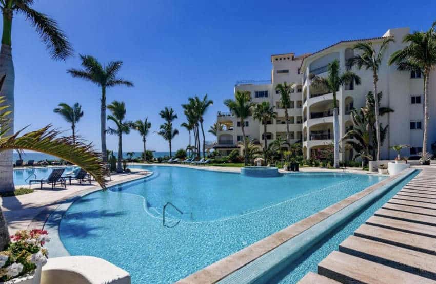 A large, curving swimming pool with clear blue water is surrounded by palm trees and a multi-story white building with balconies, all under a bright blue sky.