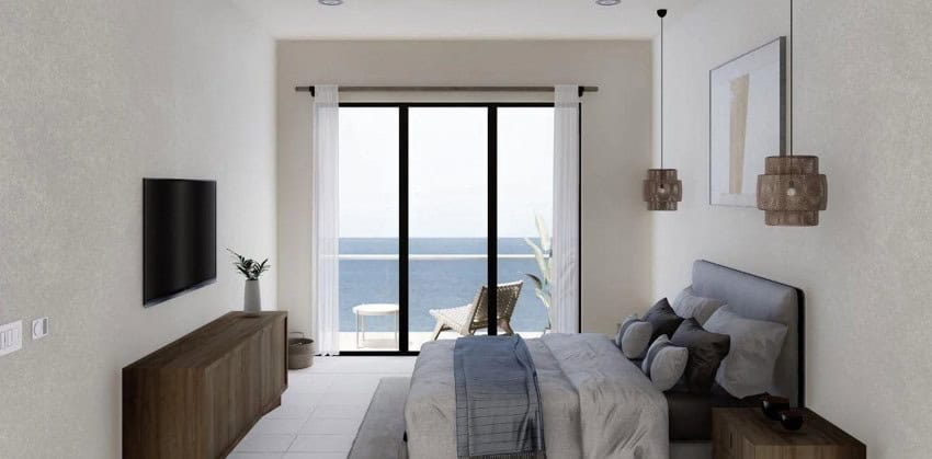 A modern bedroom with white walls features a bed with grey and blue bedding, a wall-mounted TV, and a sliding glass door leading to a balcony with an ocean view.