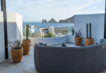A modern rooftop patio features a curved grey sofa, potted cacti, and a panoramic view of the ocean, a city, and distant rock formations under a clear sky.