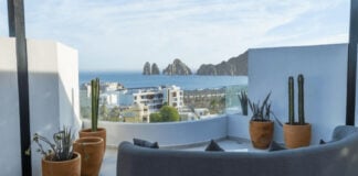 A modern rooftop patio features a curved grey sofa, potted cacti, and a panoramic view of the ocean, a city, and distant rock formations under a clear sky.