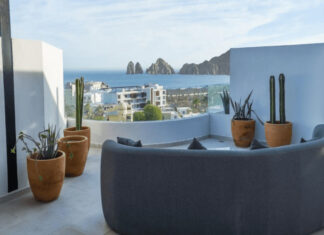 A modern rooftop patio features a curved grey sofa, potted cacti, and a panoramic view of the ocean, a city, and distant rock formations under a clear sky.