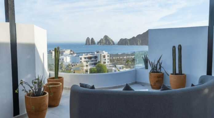 A modern rooftop patio features a curved grey sofa, potted cacti, and a panoramic view of the ocean, a city, and distant rock formations under a clear sky.