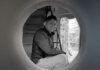 Black and white photo of artist Jaime Colín, a man with short dark hair, smiling and looking directly at the camera. He is crouched inside a large circular opening, with a staircase visible behind him and natural light illuminating a tree-like structure outside the opening.
