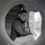 Black and white photo of artist Jaime Colín, a man with short dark hair, smiling and looking directly at the camera. He is crouched inside a large circular opening, with a staircase visible behind him and natural light illuminating a tree-like structure outside the opening.