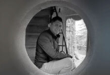 Black and white photo of artist Jaime Colín, a man with short dark hair, smiling and looking directly at the camera. He is crouched inside a large circular opening, with a staircase visible behind him and natural light illuminating a tree-like structure outside the opening.