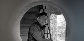 Black and white photo of artist Jaime Colín, a man with short dark hair, smiling and looking directly at the camera. He is crouched inside a large circular opening, with a staircase visible behind him and natural light illuminating a tree-like structure outside the opening.