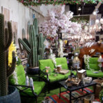 A vibrant Mexican-themed home decor display featuring large artificial cacti, bright green patio furniture, sunflowers, a cow statue, and various rustic lanterns and lighting fixtures, suggesting an eclectic outdoor living space.