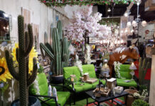 A vibrant Mexican-themed home decor display featuring large artificial cacti, bright green patio furniture, sunflowers, a cow statue, and various rustic lanterns and lighting fixtures, suggesting an eclectic outdoor living space.